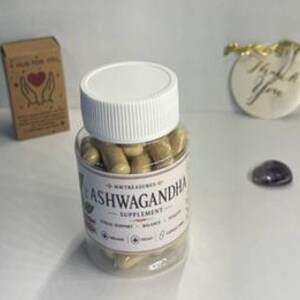 May include: A clear plastic bottle filled with tan capsules labeled "Ashwagandha Supplement." A small cardboard box with the text "A hug for you" and a "Thank You" gift tag are in the background. A small purple stone is also visible.