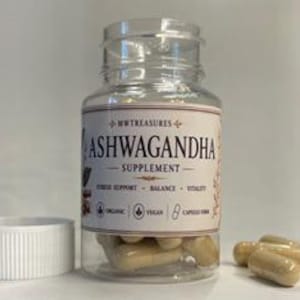 May include: A clear plastic bottle of Ashwagandha supplement capsules with a white lid. The label reads "Ashwagandha Supplement" with supporting text. Several capsules are outside the bottle. The bottle is on a white surface.