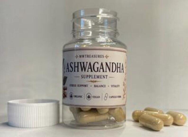 May include: A clear plastic bottle of Ashwagandha supplement capsules with a white lid. The label reads "Ashwagandha Supplement" with supporting text. Several capsules are outside the bottle. The bottle is on a white surface.