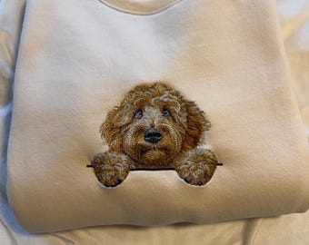 May include: A cream-colored crewneck sweatshirt featuring a detailed embroidered graphic of a brown curly-haired dog peeking over a horizontal line. The dog has expressive eyes and a black nose, with its front paws resting on the line.