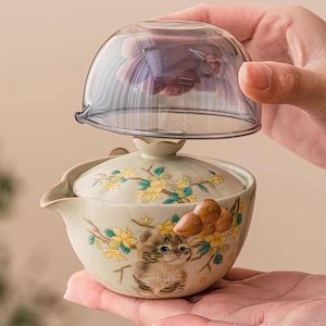 May include: A ceramic tea set with a cat design. The set includes a lidded teapot with a spout and a clear glass lid. The teapot is decorated with yellow flowers and a cartoon cat. The set is held in a person's hands.