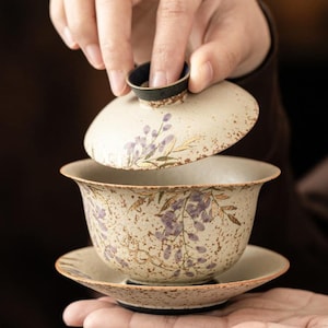 May include: A ceramic tea set with a lid, cup, and saucer. The set features a cream-colored base with purple floral designs and gold accents. The lid is being lifted, revealing the cup and saucer below. The set is designed for tea drinking.