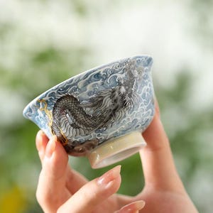 Peut inclure: Un petit bol en céramique avec un motif de dragon. Le bol présente un fond bleu clair avec une illustration détaillée d'un dragon gris. Le dragon est entouré de motifs de vagues blanches. La base du bol est de couleur jaune clair.