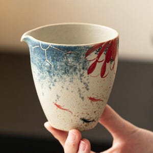 May include: A ceramic tea cup with a spout, featuring a blue and white speckled design. The cup is adorned with red floral and fish motifs. The cup is held by a person.
