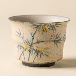以下が含まれることがあります： 竹のデザインが施されたベージュ色のセラミックティーカップ。カップには、青と金の竹の葉と茎が描かれています。カップの縁は水色で、底は濃い青色です。