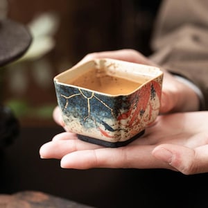May include: A square ceramic teacup with a blue and red design, filled with tea. The cup has gold accents and is held in open hands. The cup is resting on a black base.