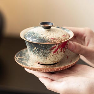 May include: A ceramic tea set with a lid, cup, and saucer. The cup and saucer have a cream-colored base with blue, red, and gold accents. The lid has a dark blue knob. The tea set is held in a person's hands.