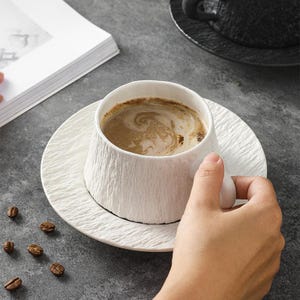 May include: White ceramic coffee cup and saucer set with a textured, bark-like design. The cup is filled with coffee, and several coffee beans are scattered around the saucer. The set is on a gray surface.