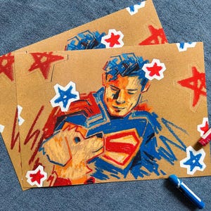 May include: Two kraft paper cards with a crayon illustration of a superhero and a dog, decorated with red and blue star stickers. A red and blue crayon and a blue crayon are on the side.