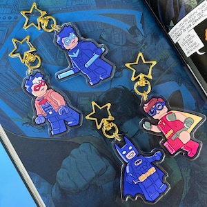 Pode incluir: Chaveiros de acrílico com personagens de super-heróis em estilo de figura de bloco. Os chaveiros têm fechos dourados em forma de estrela. Os personagens incluem Batman, Robin e Nightwing, cada um em suas cores características. Os chaveiros são exibidos em uma página de quadrinhos.