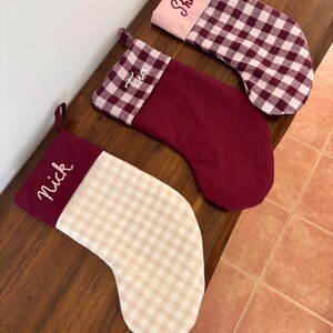 May include: Three Christmas stockings with embroidered names on the cuffs. One has a pink cuff and a white and burgundy checked pattern. Another has a burgundy cuff and body. The third has a burgundy cuff and a tan and white checked pattern.