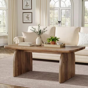 May include: A rectangular wooden coffee table with a natural finish. The table has a thick top and two arched supports. A white vase with greenery, books, and a tray with a plant and bowl are on the table. The table is in front of a cream-colored sofa.