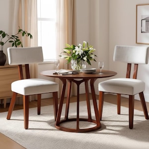 May include: A circular wooden dining table and two matching chairs. The table has a dark brown finish and a distinctive base. The chairs feature white upholstered seats and backs, with wooden frames. A vase of white flowers is on the table.