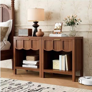 May include: Two matching wooden bedside tables with scalloped drawer fronts and open shelves. Each table has a gold-toned handle and is filled with decorative items like books, a lamp, and a vase of flowers. The tables are in a bedroom setting.