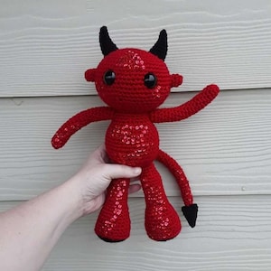 May include: A handmade red crochet devil doll with black horns, eyes, and a tail. The doll has a sequined body. The doll's arms are outstretched, and it has large, round eyes. The doll is being held up by a person's hand.