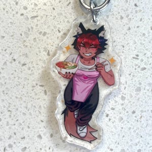May include: A clear acrylic charm featuring a cartoon character with red hair, black ears, and a pink apron. The character holds a bowl of food and chopsticks, with star accents. A silver keyring is attached.
