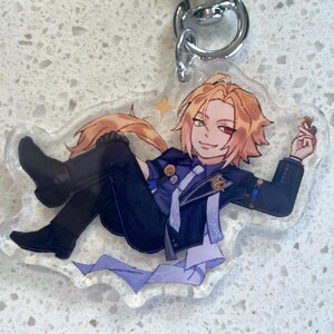 May include: A clear acrylic keychain featuring an anime-style character with blonde hair, a red and green eye, and a dark blue outfit. The character is reclining and holding a small object. The keychain has a silver metal ring.