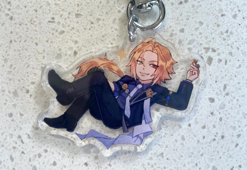 May include: A clear acrylic keychain featuring an anime-style character with blonde hair, a red and green eye, and a dark blue outfit. The character is reclining and holding a small object. The keychain has a silver metal ring.