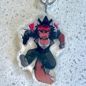 May include: A clear acrylic keychain featuring a dynamic anime-style character with red hair and a fox tail. The character is wearing a black jacket, white shirt, and dark green pants. The keychain has a silver metal ring.