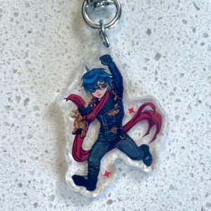 May include: A clear acrylic keychain featuring a cartoon character with blue hair, sunglasses, and a red scarf. The character is wearing a black outfit and is in a dynamic pose. The keychain has a silver metal clasp.