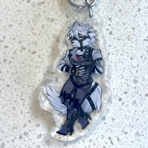 May include: A clear acrylic keychain featuring a stylized anthropomorphic wolf character in a dark outfit. The character has white fur, a black mask, and is wearing a vest with a red heart. The keychain has a silver metal ring.