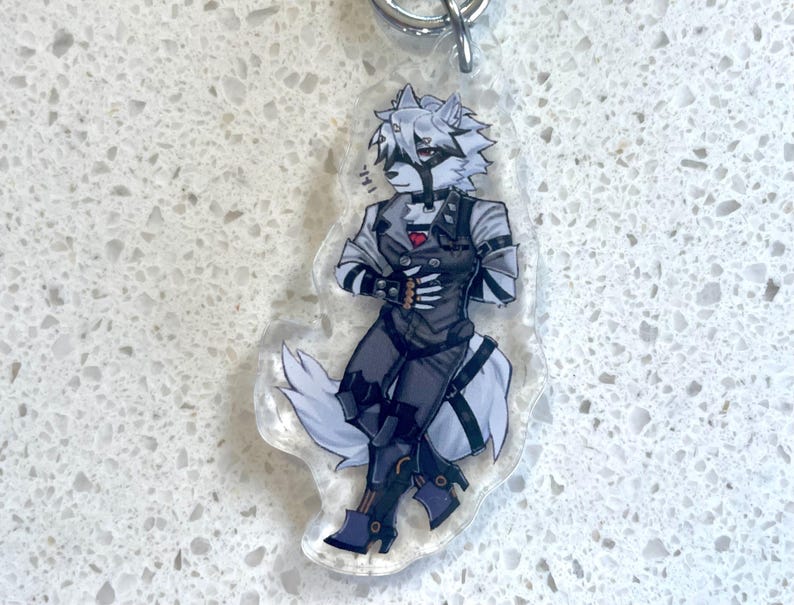 May include: A clear acrylic keychain featuring a stylized anthropomorphic wolf character in a dark outfit. The character has white fur, a black mask, and is wearing a vest with a red heart. The keychain has a silver metal ring.