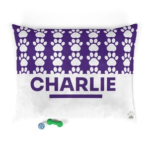 Puede incluir: Una cama rectangular para mascotas con un estampado de huellas de color púrpura en la sección superior y el nombre "CHARLIE" en letras grandes de color púrpura oscuro en la sección inferior blanca. Un pequeño juguete en forma de hueso verde y una pelota azul están frente a la cama.