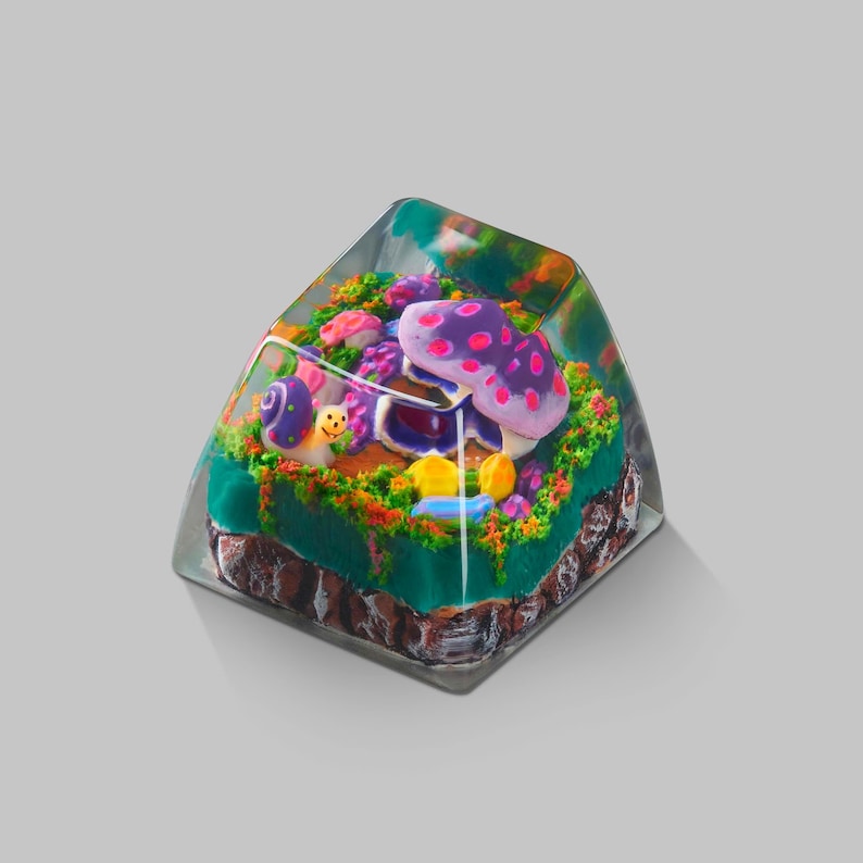 Puede incluir: Una tecla transparente y angular con una escena de fantas&iacute;a en miniatura. La escena incluye una gran seta morada con lunares rosas, un caracol y follaje colorido. La base de la tecla tiene un aspecto marr&oacute;n y texturizado.