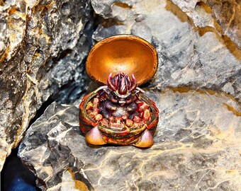 Puede incluir: Un recipiente dorado abierto con una criatura roja y dorada en su interior, posado sobre una roca gris. La criatura tiene una corona roja y está rodeada de piedras rojas y marrones. El recipiente está abierto, revelando el interior detallado.