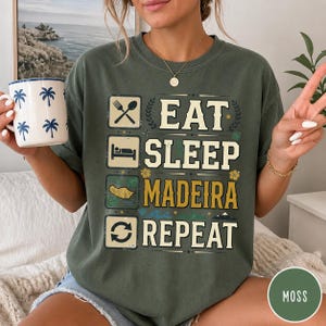 Puede incluir: Una camiseta verde musgo con un estampado gráfico con el texto EAT SLEEP MADEIRA REPEAT. El diseño incluye iconos de un tenedor y una cuchara, una persona durmiendo, un mapa de Madeira y flechas circulares, con pequeñas estrellas y flores.