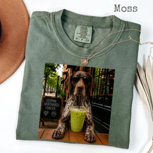 May include: A moss green crew neck t-shirt featuring a graphic print of a German Shorthaired Pointer wearing black sunglasses and drinking from a clear plastic cup with a straw. Next to the dog is a chalkboard sign that reads German Shorthaired Pointer.