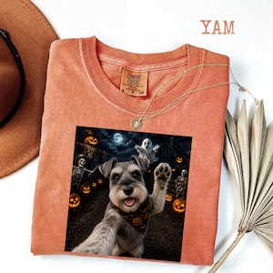 Puede incluir: Una camiseta de cuello redondo color terracota con un estampado gráfico de un perro Schnauzer que lleva un pañuelo con estampado de calabazas, rodeado de esqueletos, un fantasma y calabazas de Halloween bajo una luna llena.