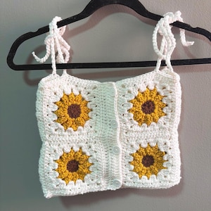 Puede incluir: Top de ganchillo blanco con diseño de girasoles. El top presenta cuatro motivos de girasoles en amarillo y marrón, con fondo blanco. Tiene tirantes ajustables con lazos y cierre de botones. El top se muestra en una percha negra.