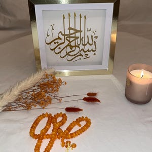 May include: A gold-framed artwork with Arabic calligraphy, a string of amber prayer beads, and a lit candle. The artwork features gold script on a white background. Dried flowers add a decorative touch.