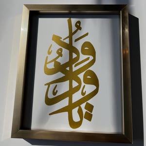 Peut inclure: Œuvre d'art encadrée avec calligraphie dorée sur fond blanc. L'écriture dorée est centrée dans un cadre rectangulaire avec une finition métallique. L'œuvre est présentée sur un mur blanc, avec des ombres ajoutant de la profondeur.