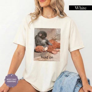 May include: Cream-colored t-shirt featuring a graphic of a monkey comforting a stuffed animal, with the words "hold on" below the image. The shirt has short sleeves and a relaxed fit.