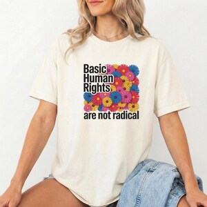 May include: A cream-coloured t-shirt featuring the text "Basic Human Rights are not radical" in black. The text is set over a square of colourful flowers in pink, yellow, blue, and red. The t-shirt has short sleeves.