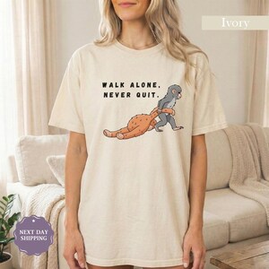 May include: Ivory-colored t-shirt with the text "WALK ALONE, NEVER QUIT." The shirt features a cartoon illustration of a gray monkey helping a brown monkey walk. The shirt is a comfortable, casual style.