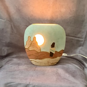 May include: A ceramic lamp with a desert landscape design. The lamp features a light blue background with a sun and a figure. The base is brown and tan. The lamp is lit from within, creating a warm glow.