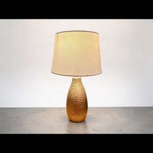 May include: A table lamp featuring a gold-colored base with a textured, hammered surface and a tapered, teardrop shape. The lamp is topped with a wide, neutral-toned fabric drum shade that provides a soft, diffused light across the room.