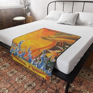 May include: A rectangular throw blanket with a vibrant design featuring a sunset, ocean waves, and tropical flowers. The blanket has the words "Eddie Aikau" in a stylized font. The color palette includes orange, yellow, blue, and white.