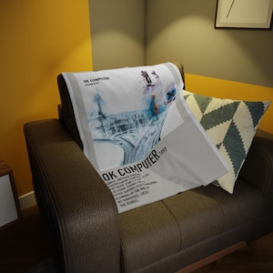 Puede incluir: Un cojín blanco con el texto "OK COMPUTER" y un diseño gráfico, apoyado en un sillón de cuero marrón. El cojín también tiene el texto "1997" y títulos de canciones. También se ve un cojín estampado.