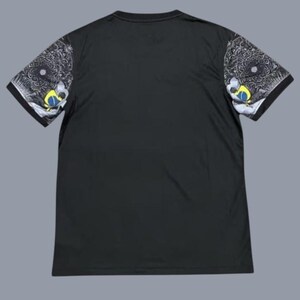 May include: Black t-shirt with short sleeves. The sleeves feature a detailed design in black, white, yellow, and blue. The design includes a circular pattern and a stylized eye. The shirt has a crew neck.