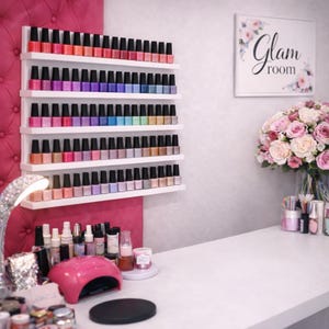 May include: A nail salon interior with a display of nail polish bottles on white shelves. The bottles are arranged in colour order. A sign that says "Glam room" is visible. A bouquet of pink roses sits on the counter. The room has a pink wall.