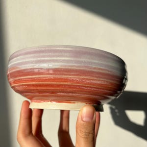 May include: A handcrafted ceramic bowl with a wide, shallow form. The bowl displays a colour gradient, shifting from a pale lavender at the top to a vivid red at the base. The bowl is held by a hand, highlighting its size and shape.