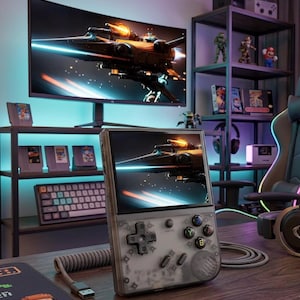 May include: A handheld gaming console with a transparent gray shell, displaying a spaceship scene. The console features a directional pad, buttons, and a screen. A large monitor behind it shows the same image. The scene includes a keyboard, game cartridges, and figurines.