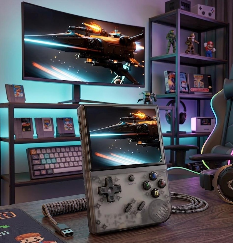 May include: A handheld gaming console with a transparent gray shell, displaying a spaceship scene. The console features a directional pad, buttons, and a screen. A large monitor behind it shows the same image. The scene includes a keyboard, game cartridges, and figurines.