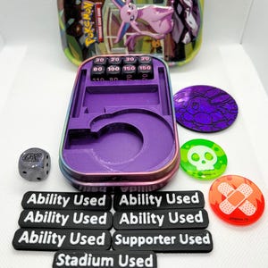 May include: A collection of Pokémon Trading Card Game accessories. Includes a metal tin, a purple damage counter, dice, tokens, and markers that read "Ability Used", "Supporter Used", and "Stadium Used".
