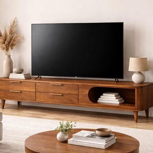 May include: A mid-century modern style wooden TV stand with drawers and an open shelf, holding books. A large flat-screen TV sits on top. The room is decorated with a lamp, plants, and a round coffee table.