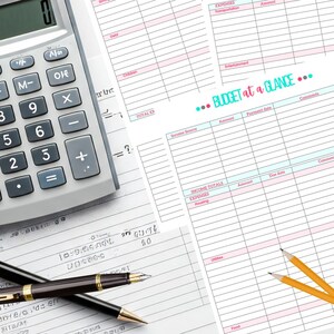 May include: A grey calculator with black buttons and a digital display is next to budget planning sheets. Two pens and two pencils are also on the white surface. The budget sheets have pink and blue lines and the text "BUDGET at a GLANCE".